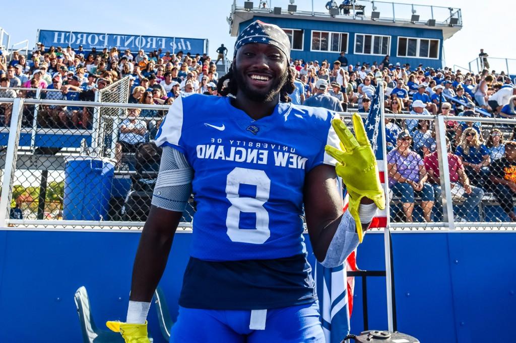 A football player smiles at the camera while standing in front of a crowd at a game